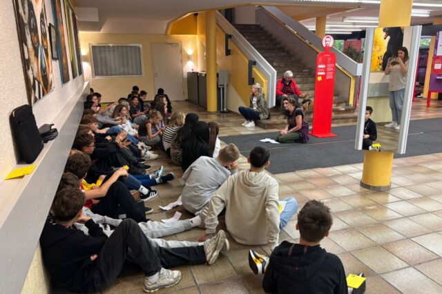 mehrere Jugendliche am Boden eines hallenartigen Raumes sitzen, einer ebenfalls am Boden sitzenden Frau zuhörend
